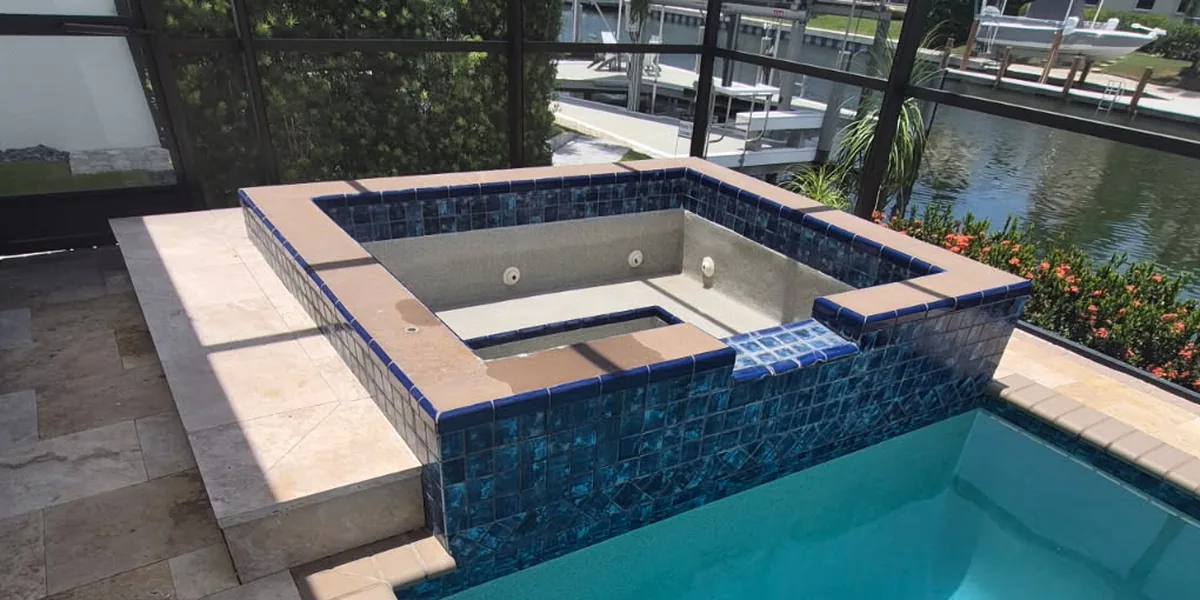 Before: a raised spa beside a canal-front screened pool on Marco Island, drained to its bare shell, with dark blue square tile on its outer face and tan brick coping around the rim